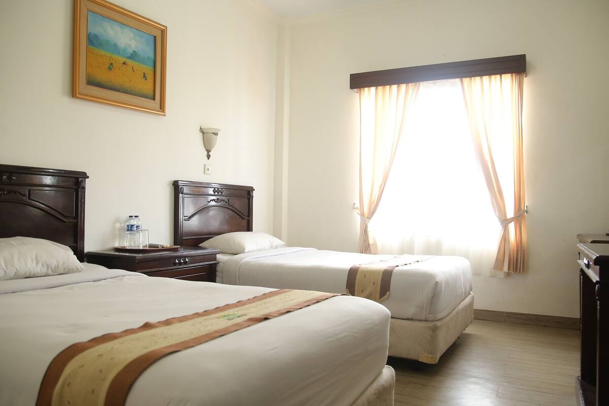 Habitación de hotel con dos camas, una mesita de noche con lámpara, un cuadro en la pared y una ventana con cortinas.