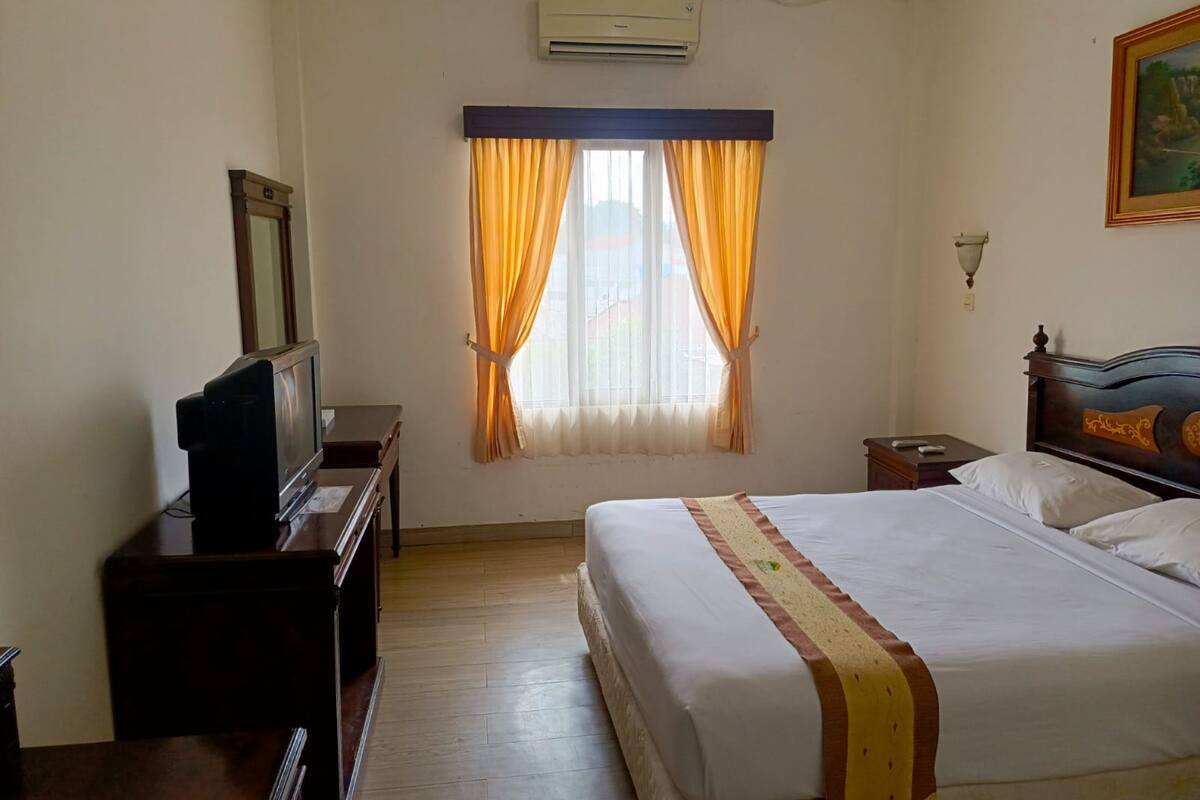 Una habitación de hotel con una cama, un televisor sobre un soporte de madera, una ventana con cortinas, un equipo de aire acondicionado y una lámpara fijada a la pared.