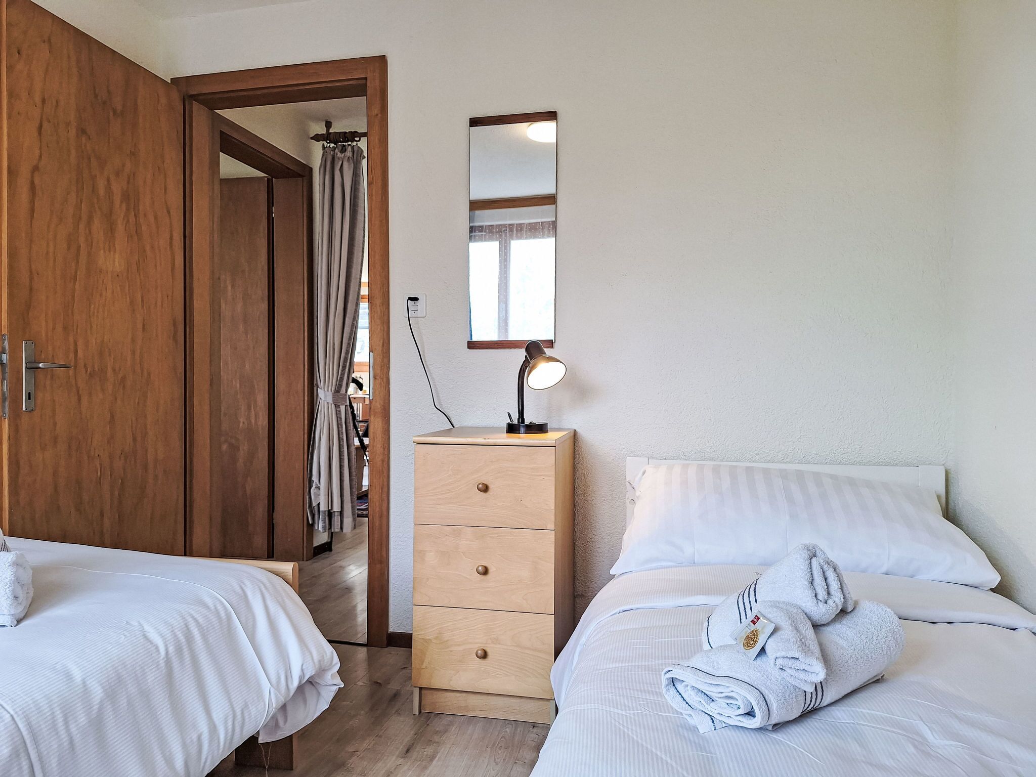 A bedroom with two beds, a wooden wardrobe, a bedside table with a lamp, and a mirror on the wall.