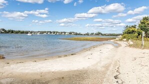 A sandy beach with a calm lake, boats, and a distant shoreline with houses. - Perfect vacation home : Cape Cod Charm & Comfort. Walk to private beach (Bourne)