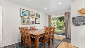 A dining area with a wooden table and chairs, framed artwork on the wall, and a view of greenery through a window. - The Abbey - Point Lonsdale (Point Lonsdale)