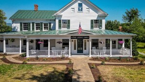 A two-story white building with a green roof, a porch with white railings, and an American flag. - The Legacy Suites Collection Full Property Rental (Luray)