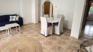 Een woonkamer met een donkerblauwe bank, een witte salontafel, een ronde eettafel met vier stoelen, een geweven vloerkleed en een rieten fauteuil met een kussen.