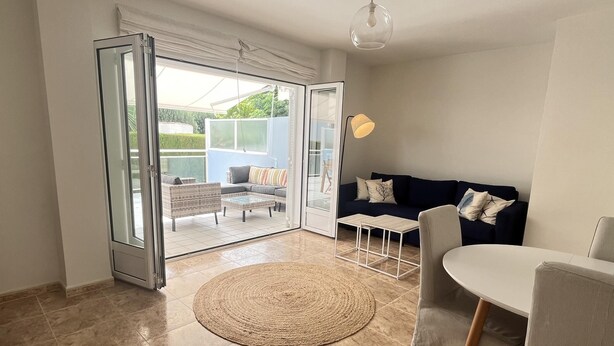 Een moderne woonkamer met een schuifdeur naar een balkon, een rond geweven vloerkleed, een donkerblauwe bank, een witte tafel en een hanglamp.