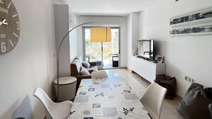 A modern living room with a dining area, a TV, and a large clock on the wall. - Flat Aguamar, ground floor, 1 bedroom, garden and swimming pool. (Dénia)