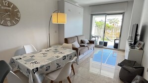 A modern living room with a dining area, a sofa, a TV, and a large window. - Flat Aguamar, ground floor, 1 bedroom, garden and swimming pool. (Dénia)