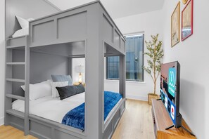 A modern bedroom with a bunk bed, a bedside table with a lamp, a TV, and a window with a view of the outside.