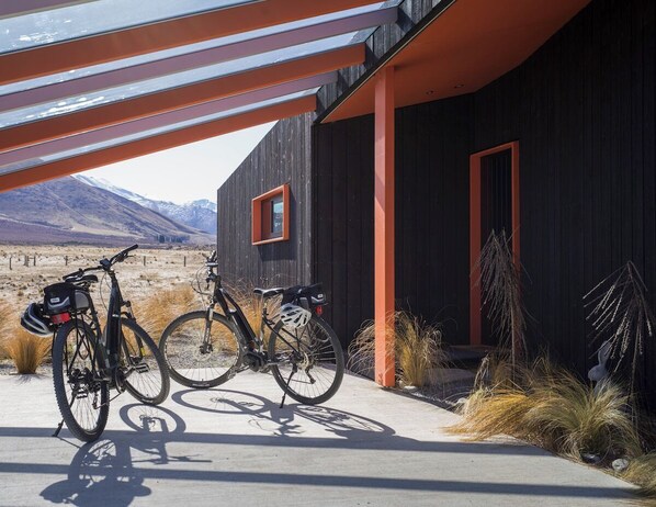 Un edificio moderno con fachada oscura y detalles naranja, dos bicicletas estacionadas afuera, y un paisaje montañoso al fondo.