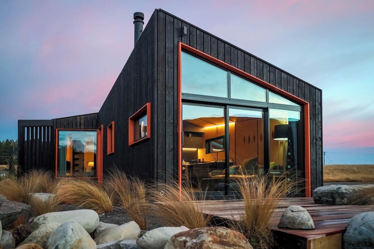 A modern, dark-colored cabin with large glass windows, a wooden deck, and a natural landscape backdrop.