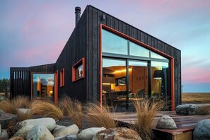 Una cabaña moderna de color oscuro con amplios ventanales, una terraza de madera y un paisaje natural de fondo.