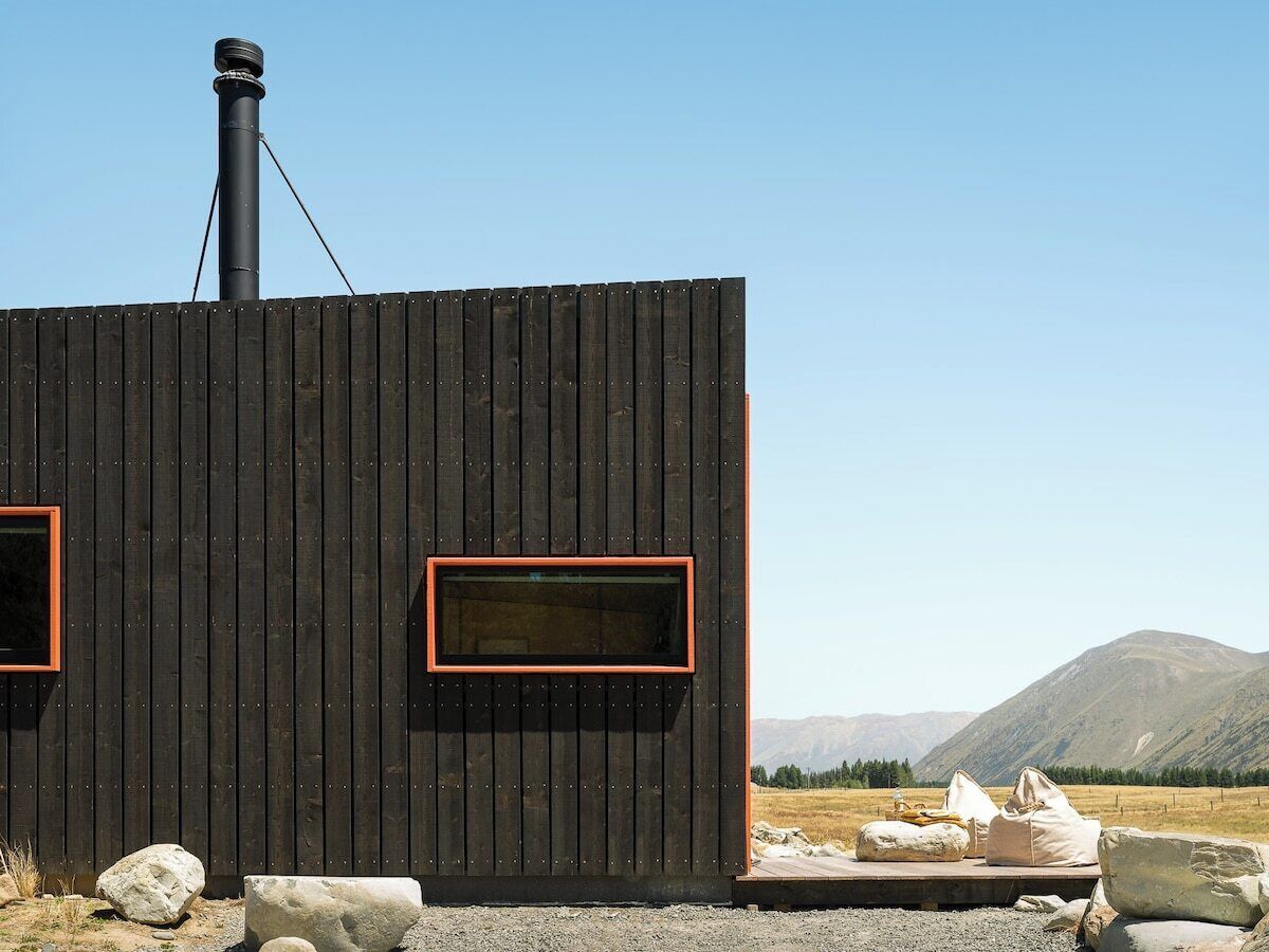 A cabin with a chimney, two windows with red frames, and a view of mountains in the background.