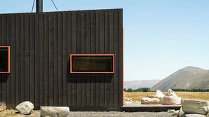 A cabin with a chimney, two windows with red frames, and a view of mountains in the background. - Skylark Cabin – Private Luxury Escape with Hot Tub (Ben Ohau)