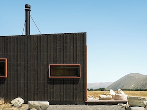 A cabin with a chimney, two windows with red frames, and a view of mountains in the background. - Skylark Cabin – Private Luxury Escape with Hot Tub (Ben Ohau)