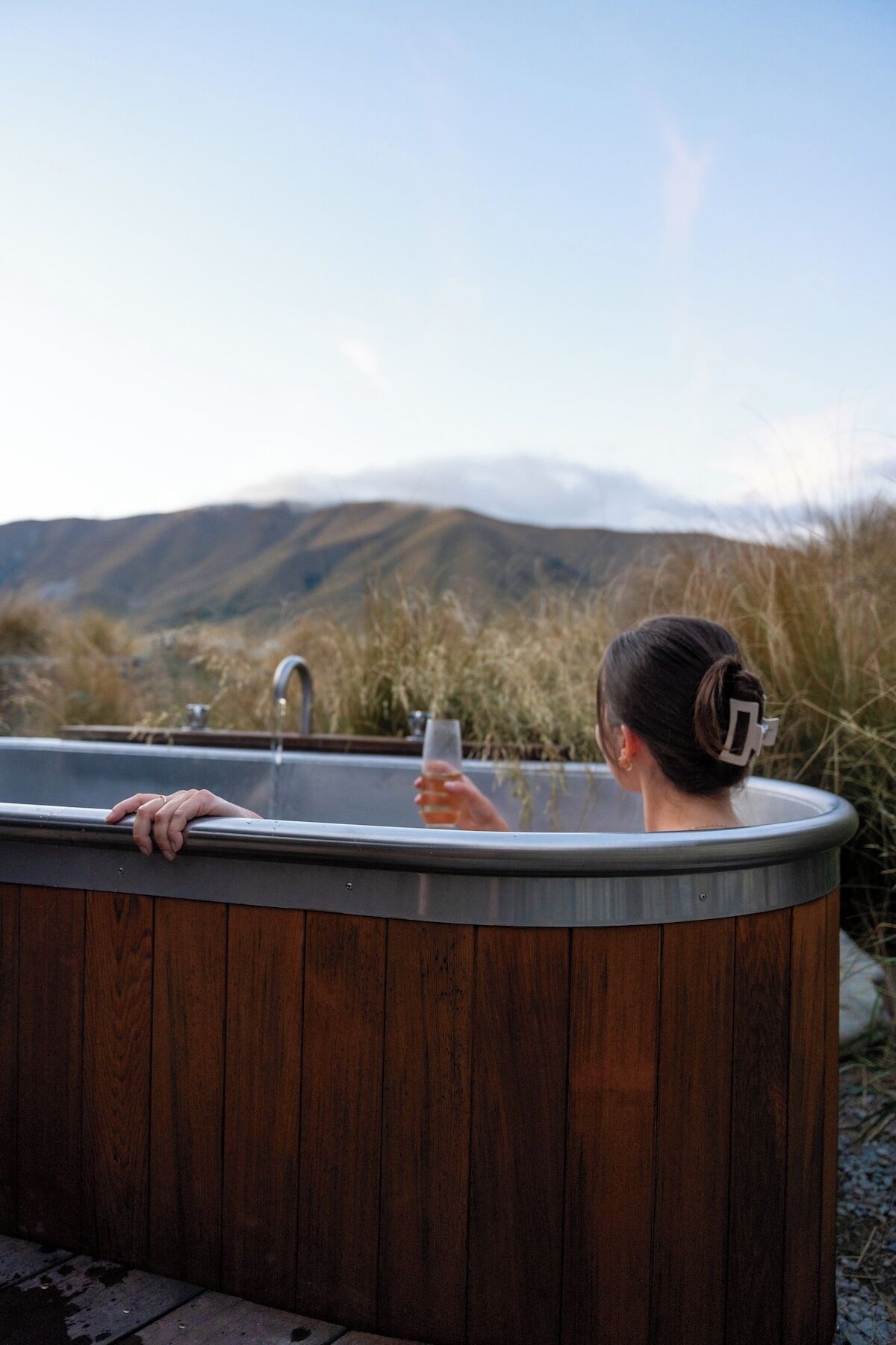 A hot tub with a wooden exterior, a metal faucet, and a person holding a glass.