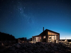 Una casa moderna con un amplio ventanal, rodeada de rocas, bajo un cielo estrellado.