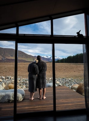 A couple standing on a wooden deck, looking out at a mountainous landscape through large windows. - Skylark Cabin – Private Luxury Escape with Hot Tub (Ben Ohau)