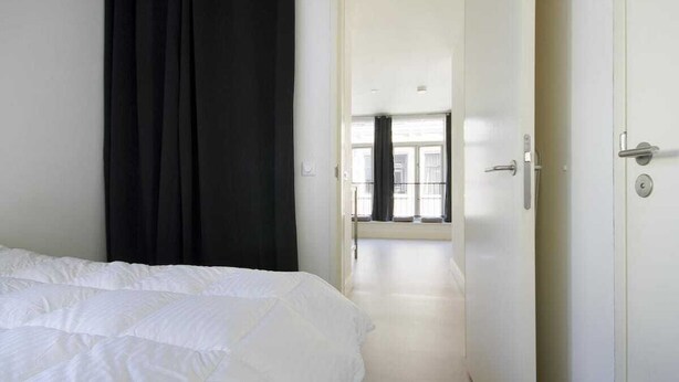 A hotel room with a bed, a door leading to a hallway, and a window with curtains.