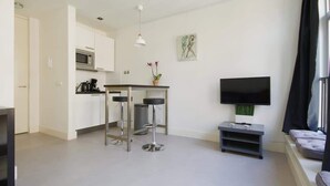 A modern living room with a flat-screen TV, a small kitchen area with a microwave, and two bar stools.