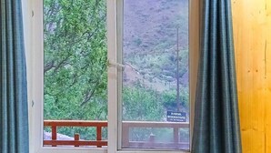 A room with a large window offering a view of a forested hillside. There are two wooden chairs and a small table with a plant and a remote control.
