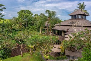 Un resort tropical con un edificio principal de techo de paja, un área de estar al aire libre cubierta y exuberante vegetación alrededor de la propiedad.