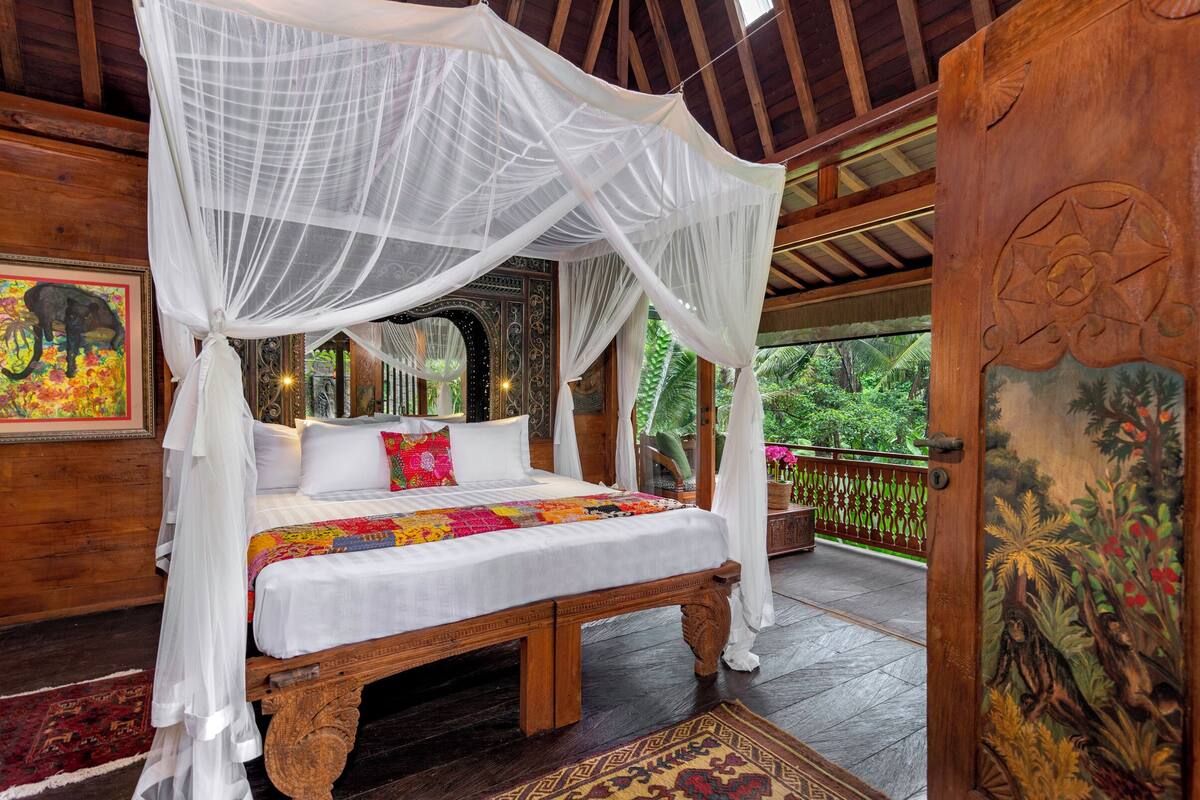 A four-poster canopy bed with a colorful quilt, a wooden floor, and a view of lush greenery outside.
