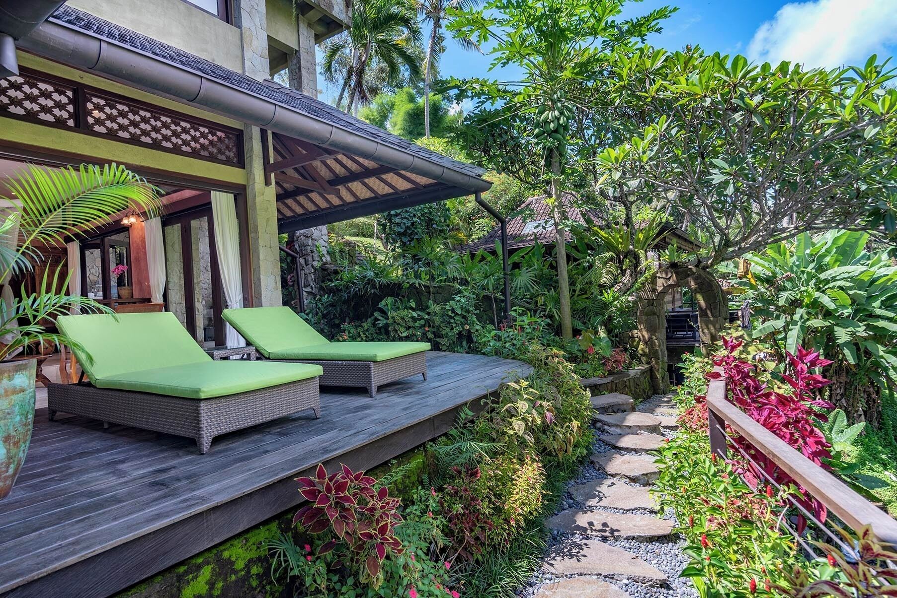 A wooden deck with two lounge chairs, surrounded by lush greenery and a stone pathway.