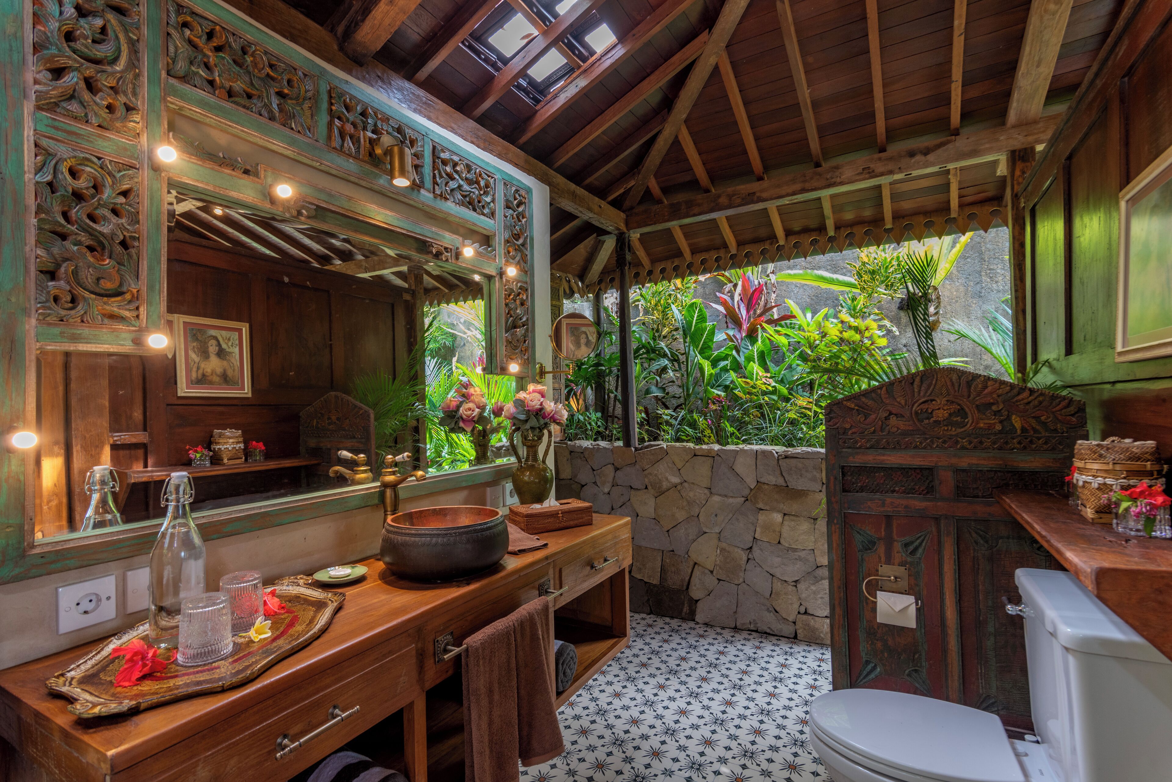 Une salle de bain avec un meuble-lavabo en bois, un grand miroir, un mur en pierre et une vue sur une végétation luxuriante à l’extérieur.