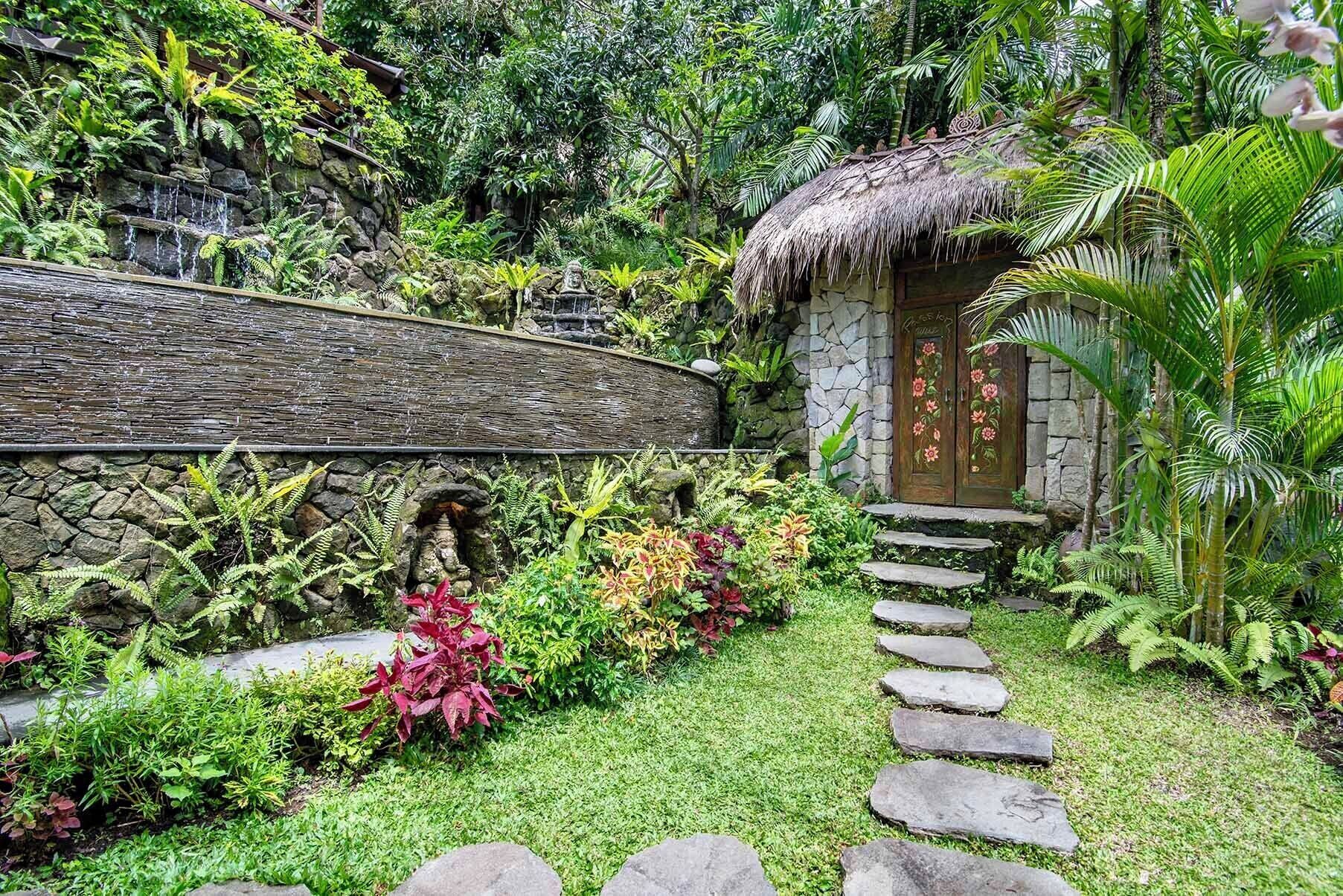 A garden pathway leading to a thatched-roof structure with a stone wall and lush vegetation.