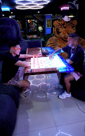 To personer sidder ved et bord med en glødende overflade, måske et spillebord, i et rum med en stor, blød tigerskulptur og en lampe i loftet.