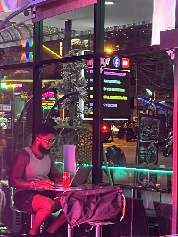 En person sidder ved et bord med en bærbar computer, en drik og en taske, i en bygning med store vinduer og neonskilte.