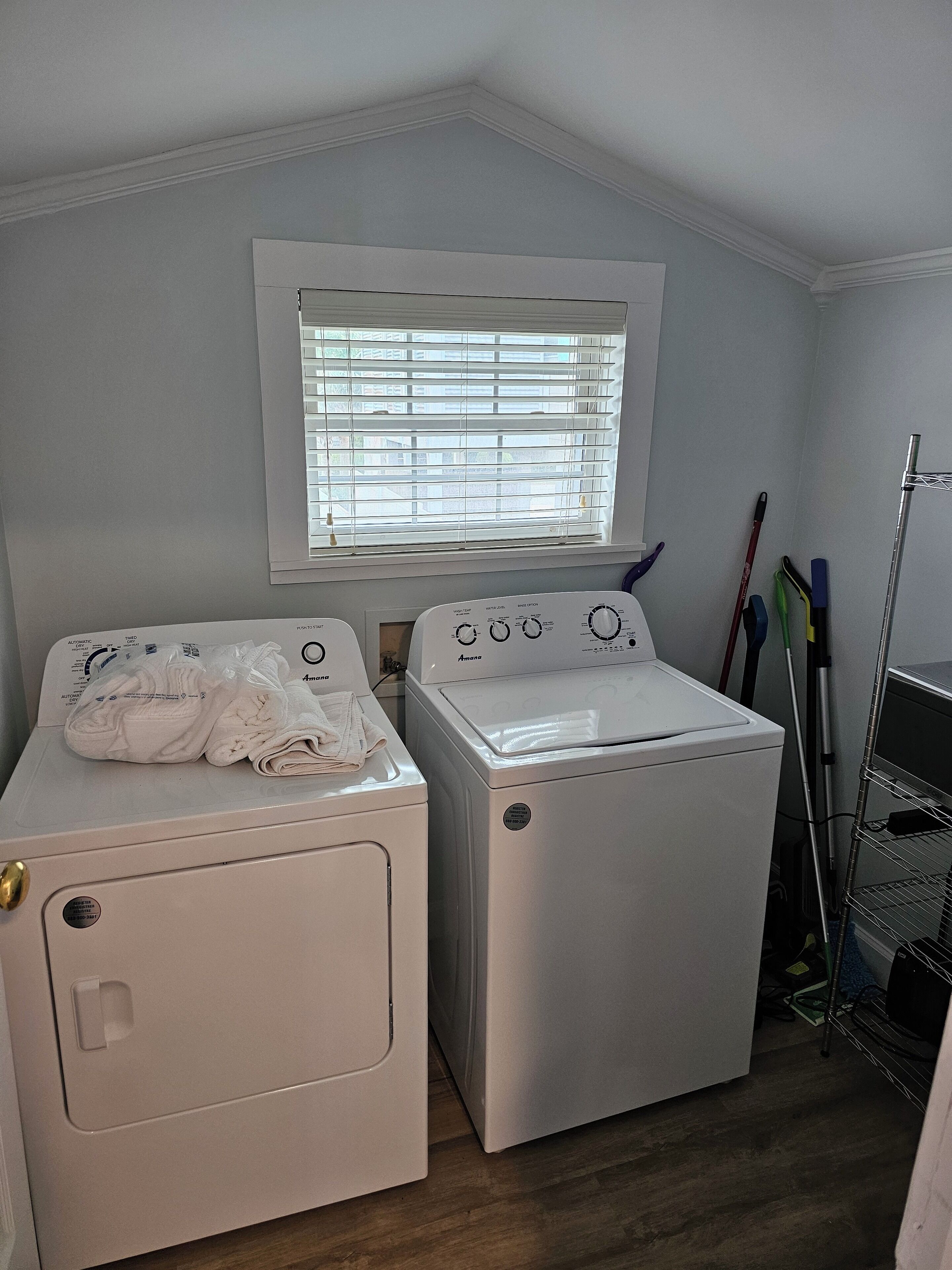 A laundry room with two front-loading washing machines, a window with blinds, and a metal rack with cleaning tools.