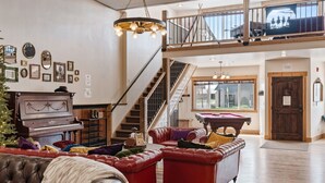 A spacious living room with a grand piano, red leather sofas, a pool table, and a staircase leading to an upper level. - Historic Yellowstone Saloon |Sleeps 25, Hot Tub, Pool Table & More! (Ashton)