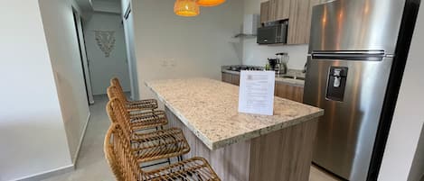 A modern kitchen with a central island, stainless steel appliances, wicker bar stools, and pendant lighting.