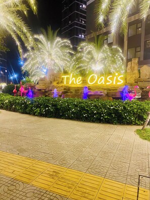 A nighttime outdoor area with illuminated text reading "The Oasis," surrounded by decorative plants and a stone wall. - Vinhomes Grand Park Luxury Apartments  (Ho Chi Minh City)