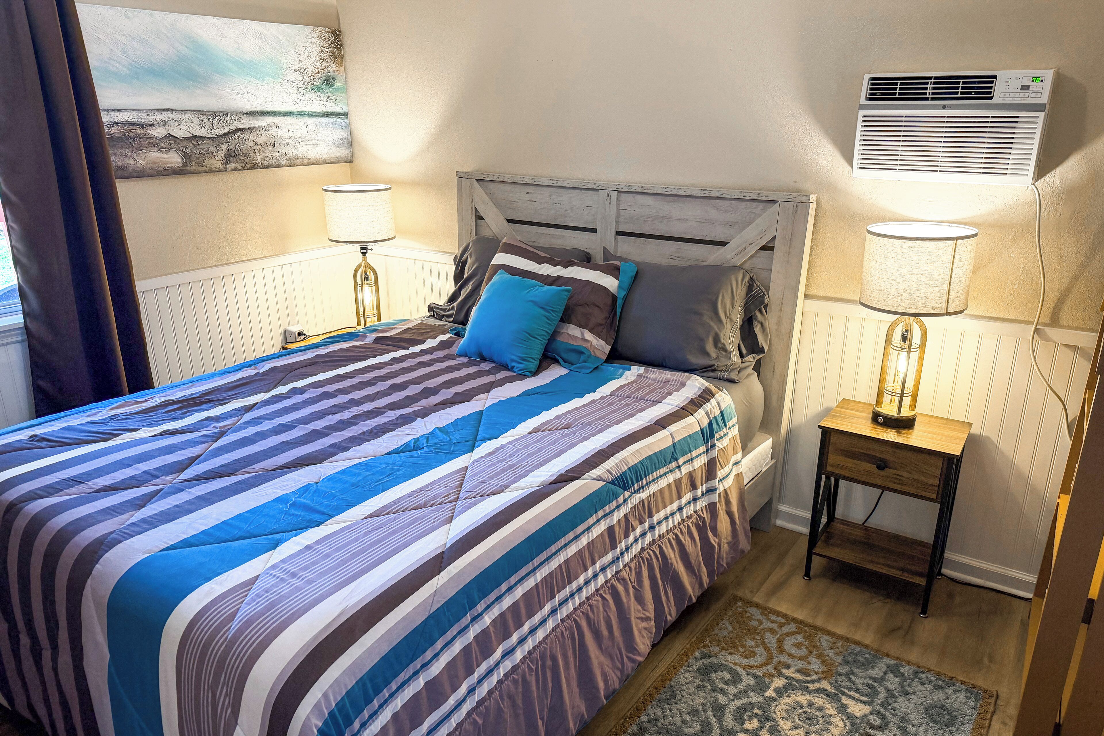 A bedroom with a bed, a nightstand, a lamp, a wall-mounted air conditioner, and a painting on the wall.