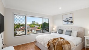 Un dormitorio con una cama grande, televisión y ventana con vista a casas.