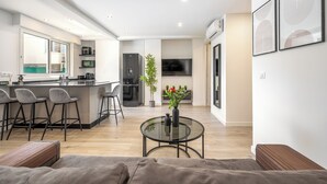 Eine moderne Küche mit Barhocker, ein Essbereich mit Glastisch und ein Wohnzimmer mit Sofa und Couchtisch.