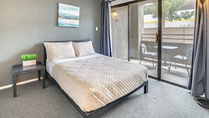 A bedroom with a bed, a nightstand with a plant, a painting on the wall, and a view of the outside balcony.