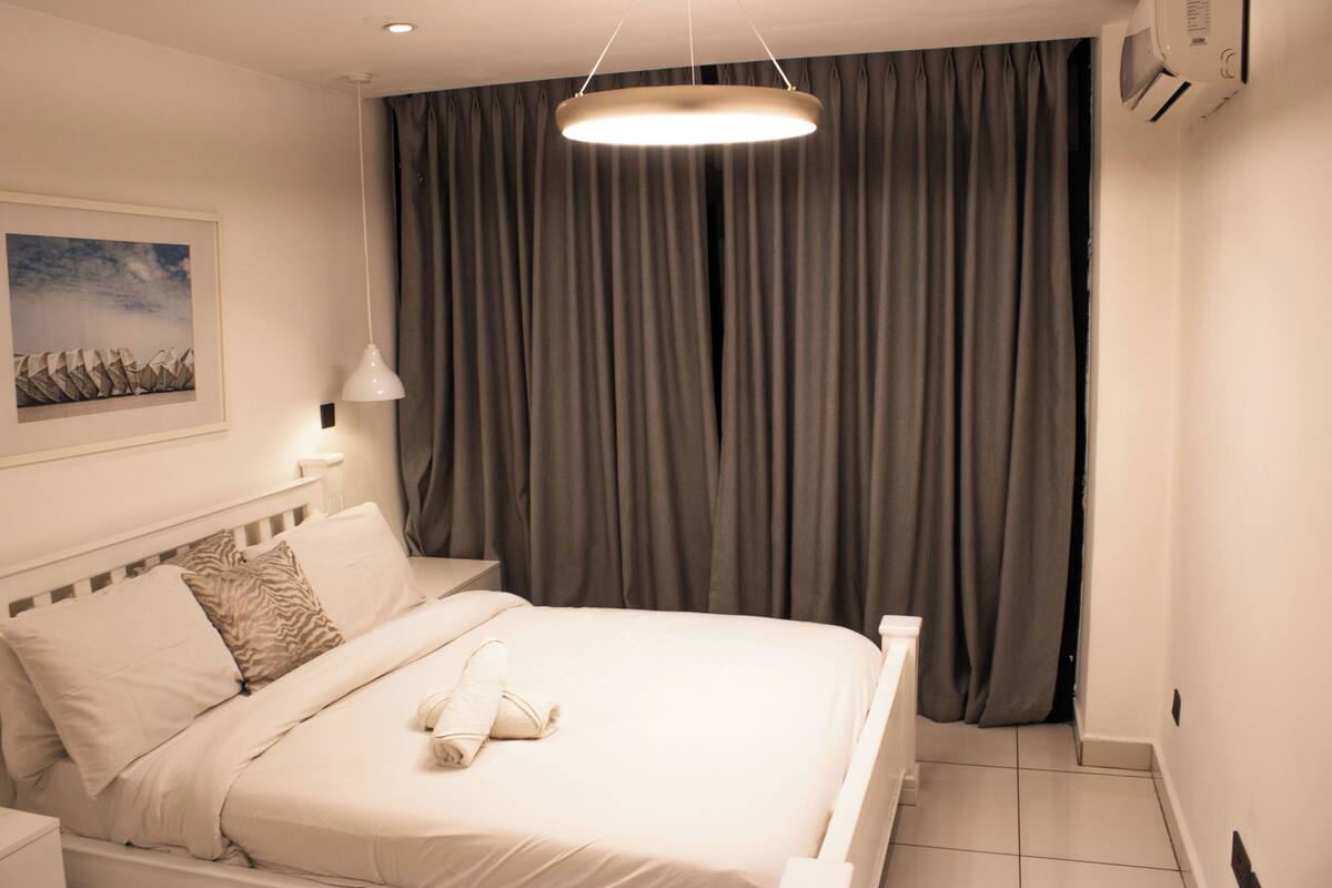 Ein modernes Hotelzimmer mit einem ordentlich bezogenen Bett, einem großen Fenster mit Vorhängen, einer wandmontierten Klimaanlage und einem gerahmten Bild an der Wand.