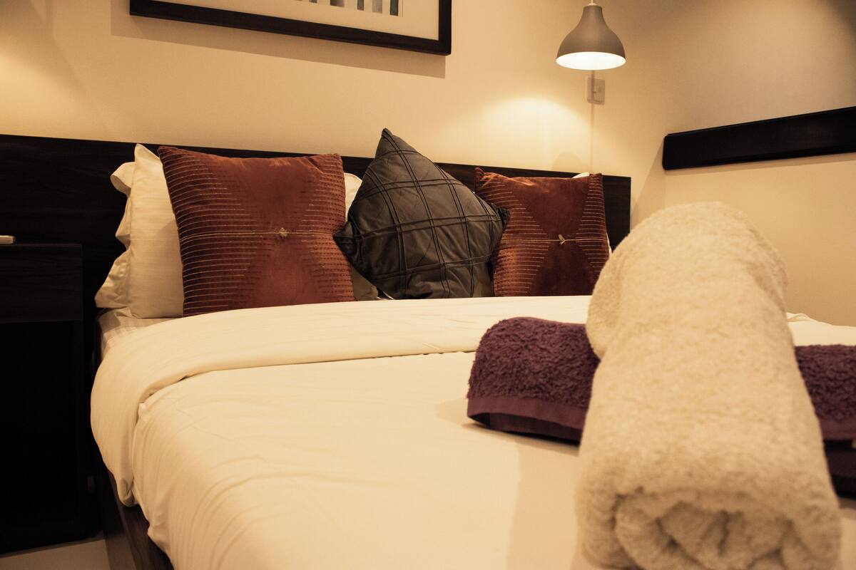 Ein ordentlich bezogenes Bett mit weißer Decke und mehreren Kissen, darunter eines mit Rastermuster in Schwarz.
