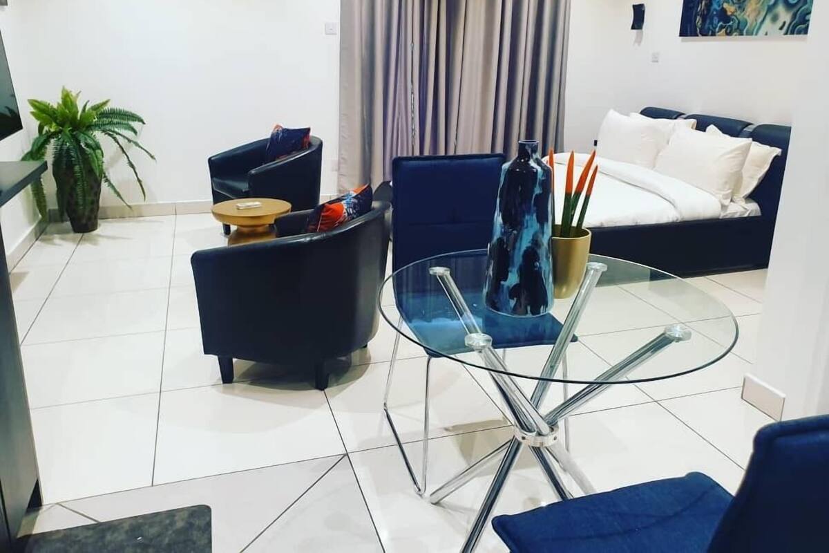 Ein modernes Wohnzimmer mit einem Glastisch, schwarzen Sofas, einem blauen Akzentstuhl, einer Topfpflanze und einem Bett mit weißer Bettwäsche.