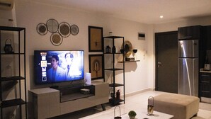 En moderne stue med et fladskærms-tv, en sofa, et sofabord og et køkkenområde med et køleskab.