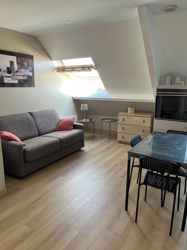 Un espace de vie compact comprenant une chambre en mezzanine, un coin cuisine, un canapé gris avec des coussins rouges, une petite table ronde et une table à manger avec des chaises.