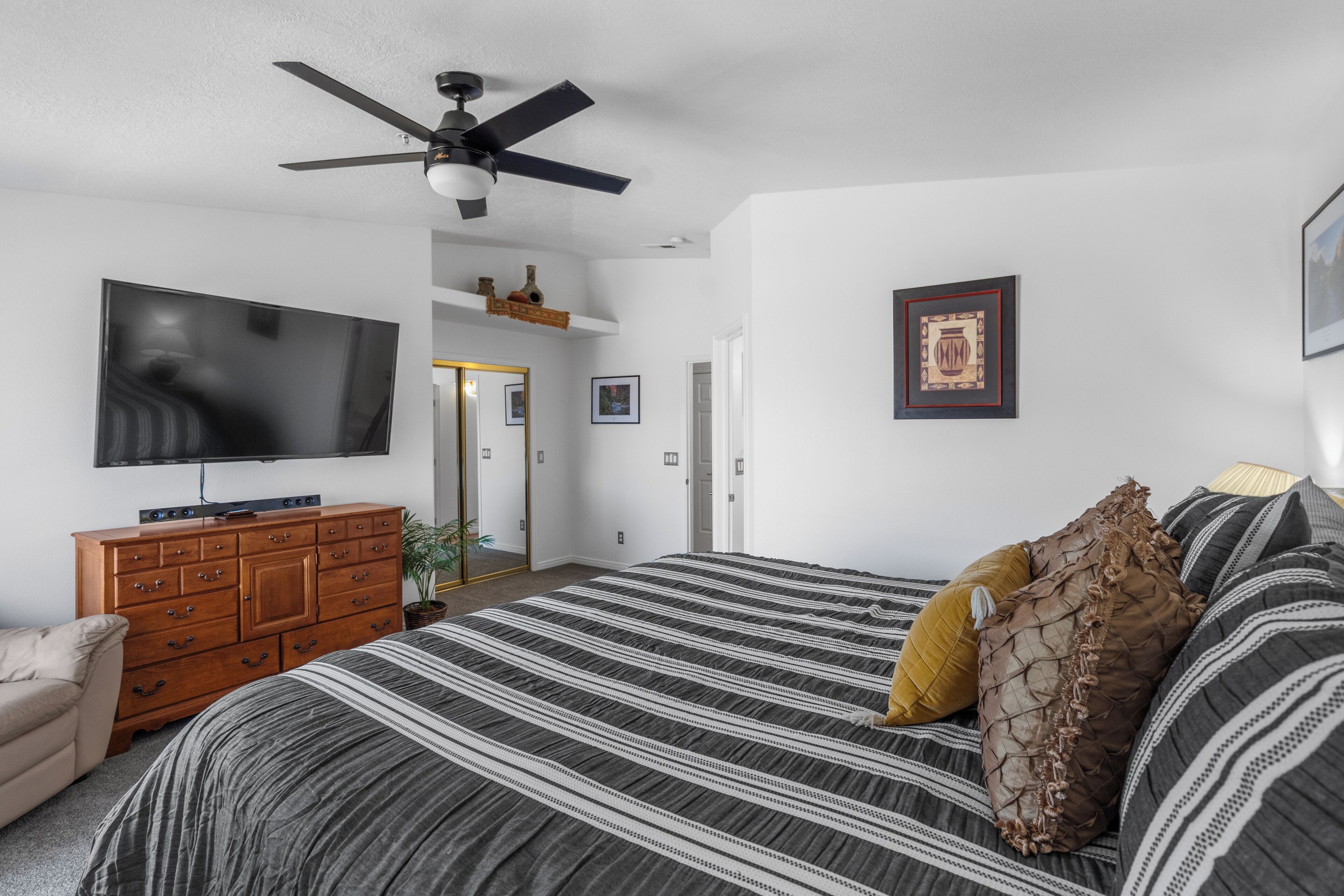A bedroom with a bed, dresser, TV, ceiling fan, and framed pictures on the wall.