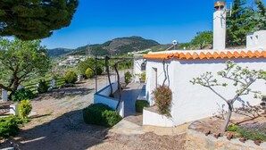 Un edificio blanco con tejas anaranjadas, un jardín con diversas plantas y una vista de un pueblo y colinas al fondo.