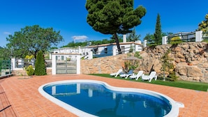Una piscina con borde de piedra, rodeada por un patio con sillones de descanso y un pequeño edificio con una verja.