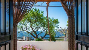 Vista a través de un marco de madera con cortina estampada, que da a un balcón con un arbusto de flores rosas en maceta y vistas de un árbol y montañas.