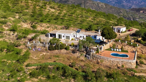 Propiedad en una ladera con piscina, un edificio blanco de tejado a la vista y una vista de montañas nevadas al fondo.