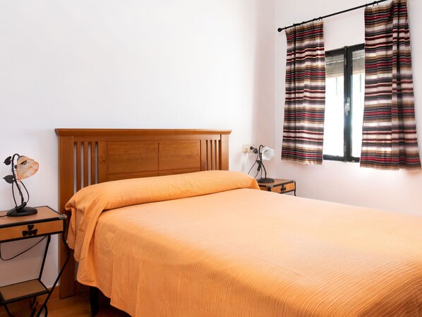 A bedroom with a wooden headboard, an orange bedspread, a nightstand with a lamp, and a window with striped curtains.