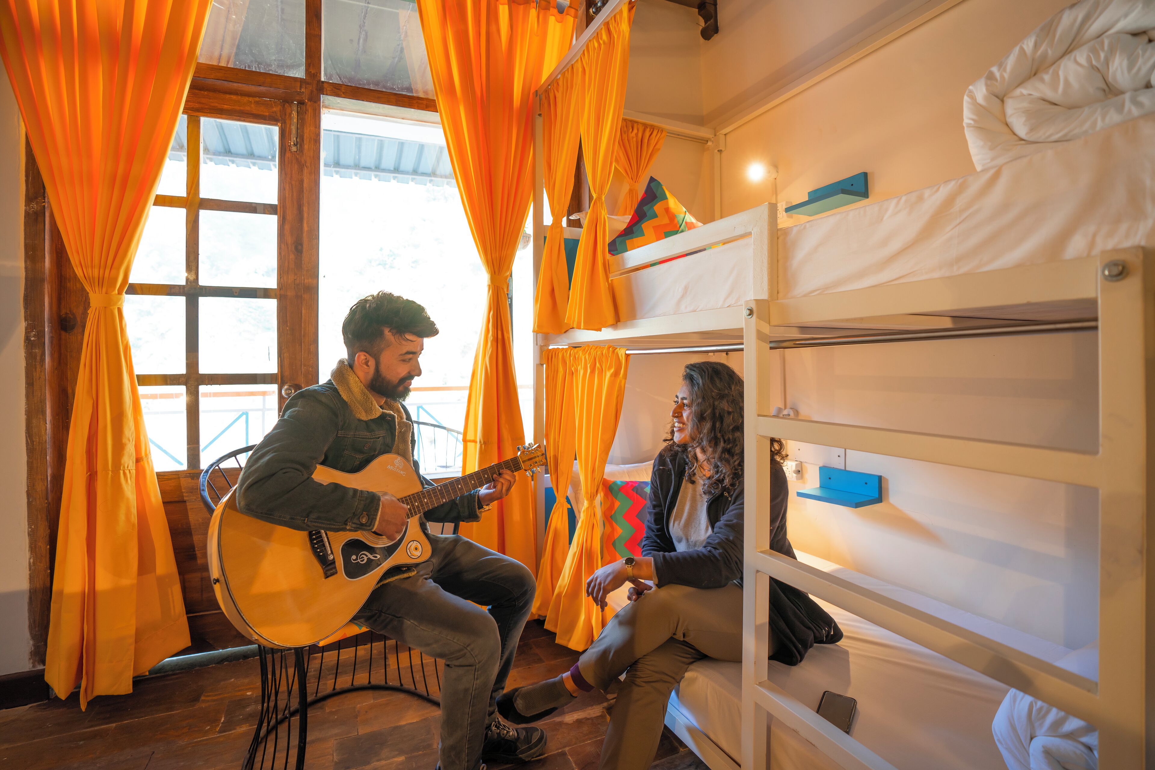 A bunk bed room with orange curtains, a guitar player, and a person sitting on the bed.
